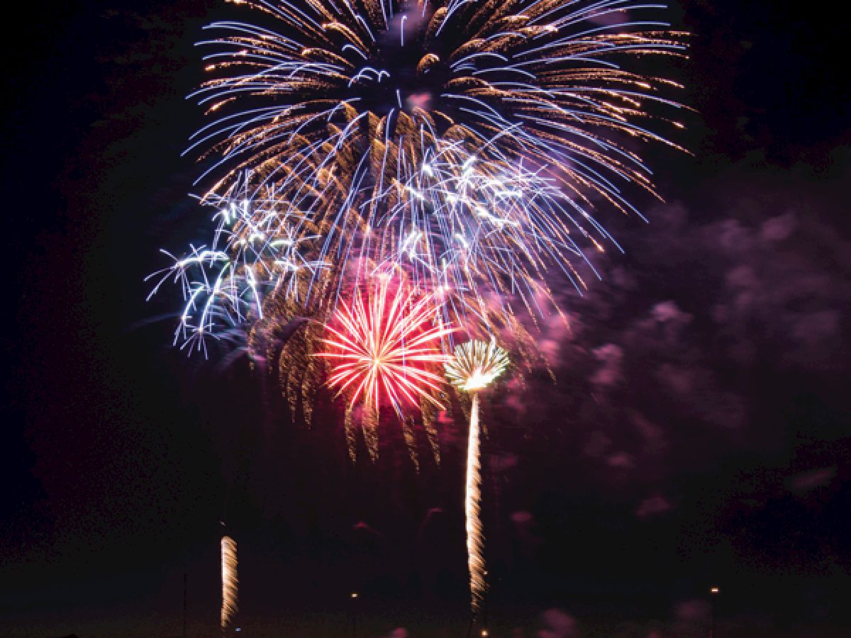 A display of colorful fireworks bursts in the night sky above a body of water, with reflections shimmering on the surface.