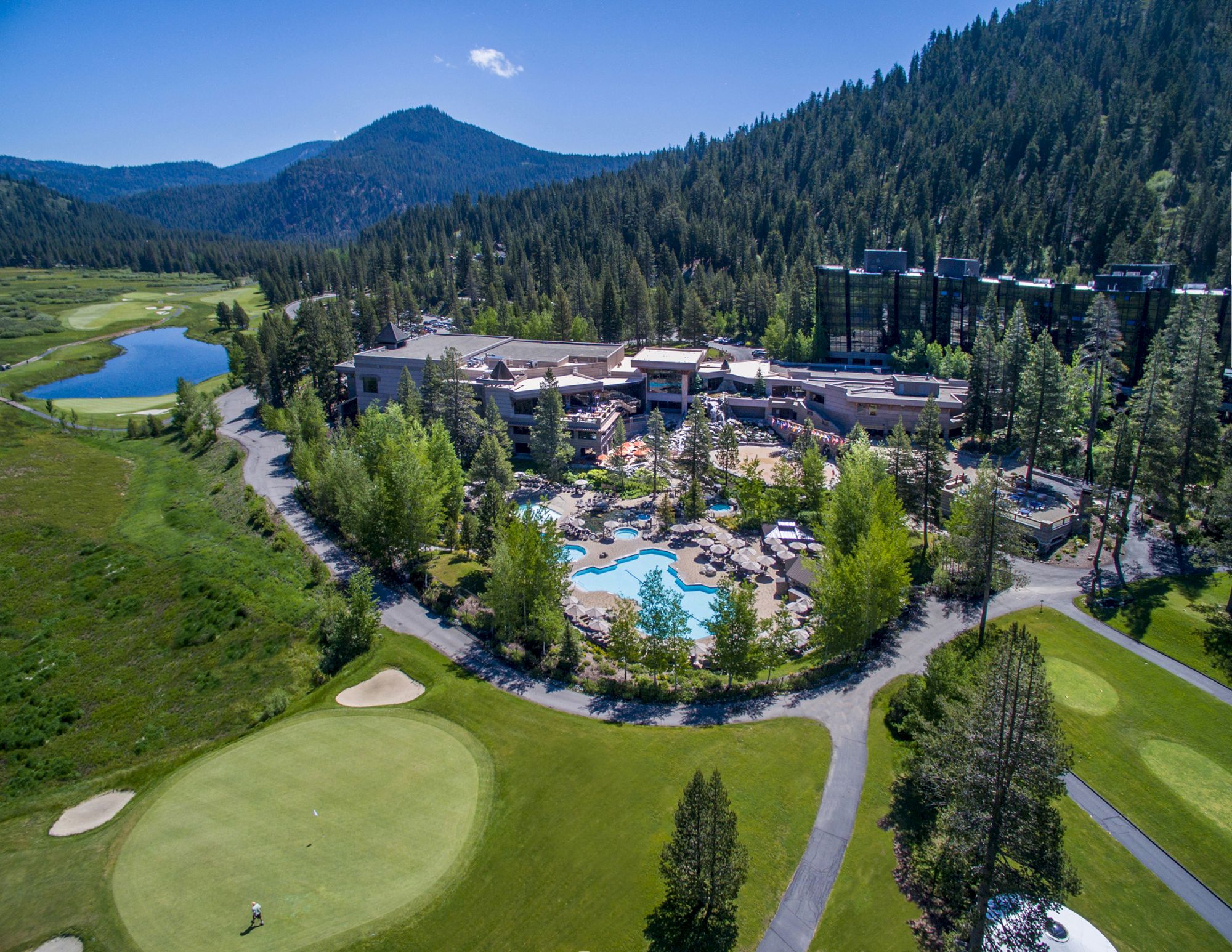 Aerial view of a resort surrounded by trees, featuring a pool area and golf course in a mountainous landscape on a clear day.
