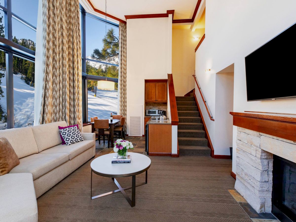 The image shows a cozy living room with a sofa, table, fireplace, TV, staircase, and large windows with a snowy outside view.