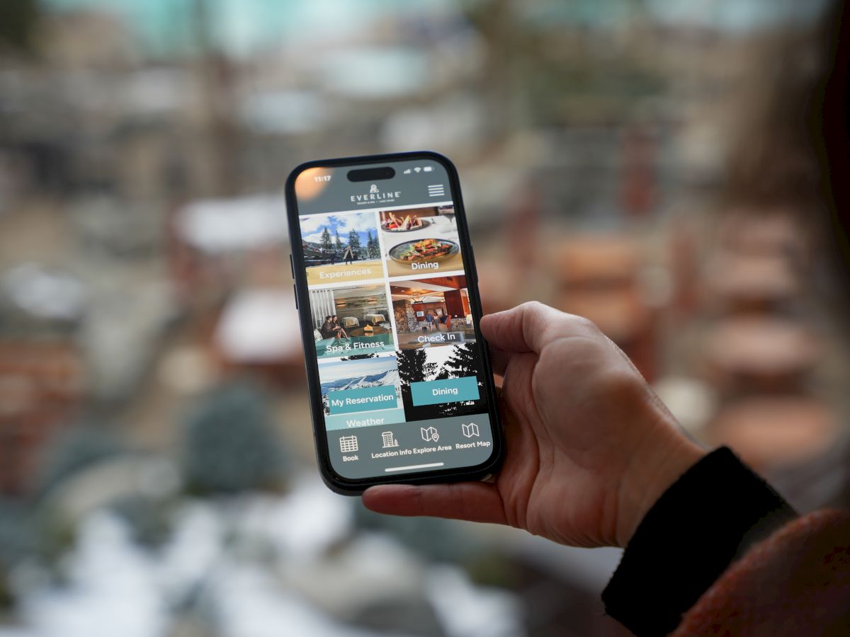 A person holding a smartphone displaying images of a resort or hotel app interface, with a blurred outdoor background in the distance.