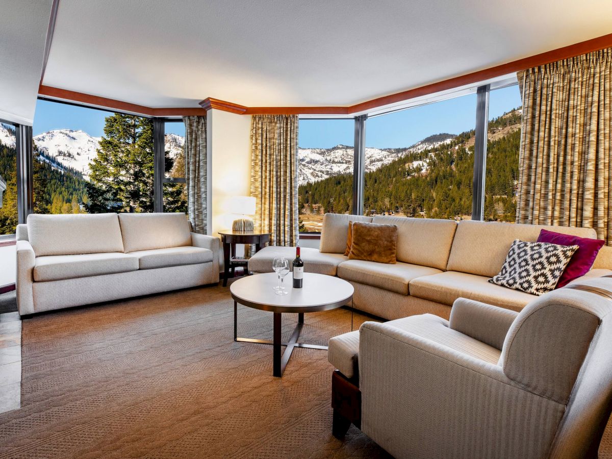 A cozy living room with sofas, armchairs, a round table, and large windows showing a scenic mountain view.