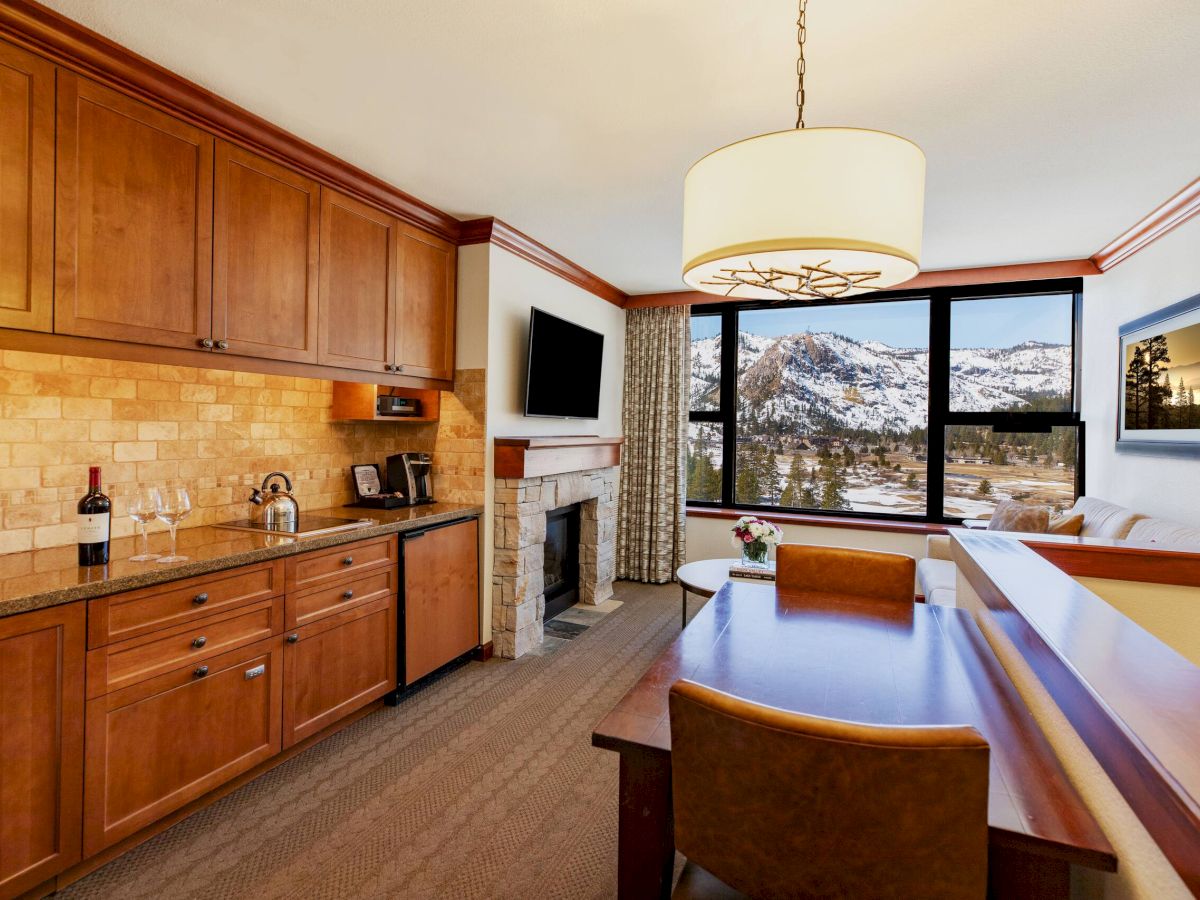 The image shows a cozy hotel room with a kitchenette, dining table, fireplace, and large window offering a scenic mountain view.