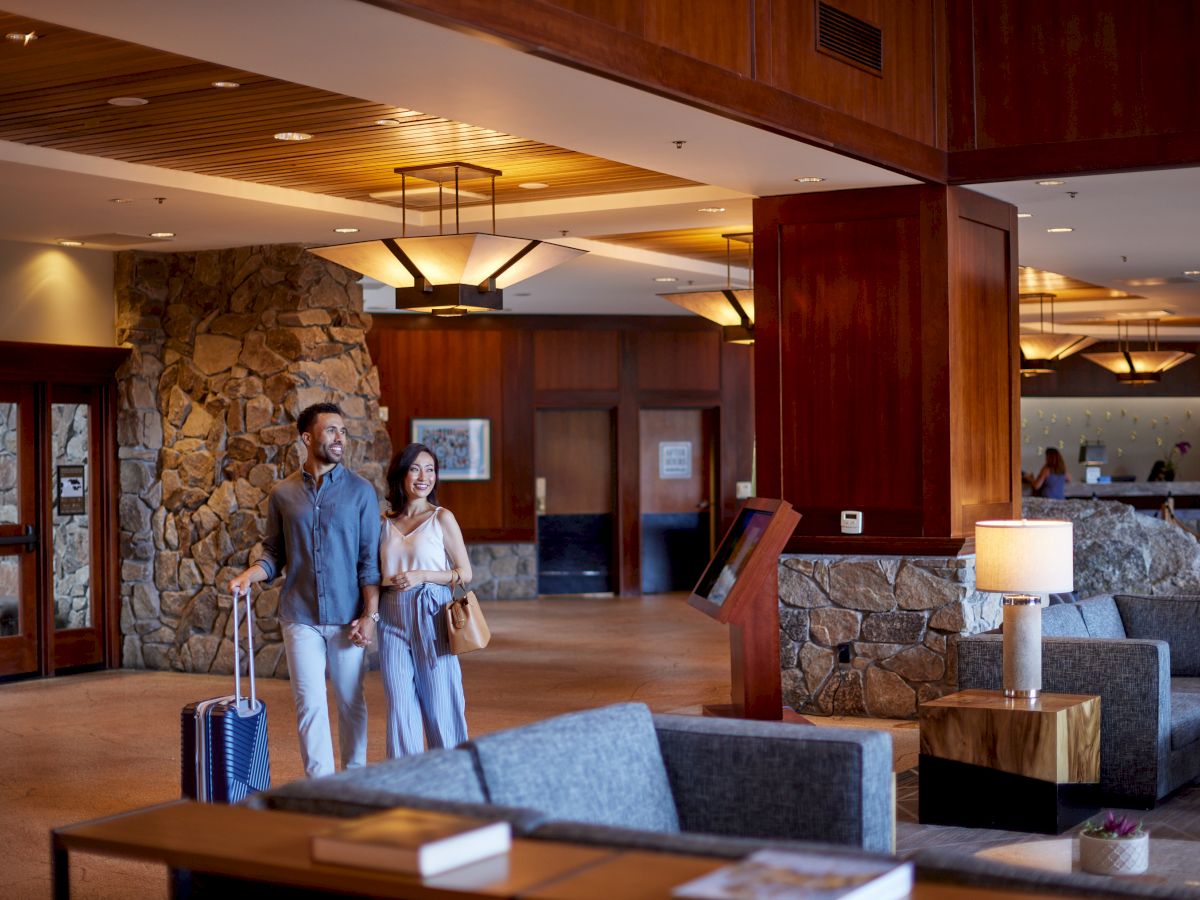 A couple walks through a stylish hotel lobby with luggage, featuring stone walls, wood finishes, and cozy seating areas.