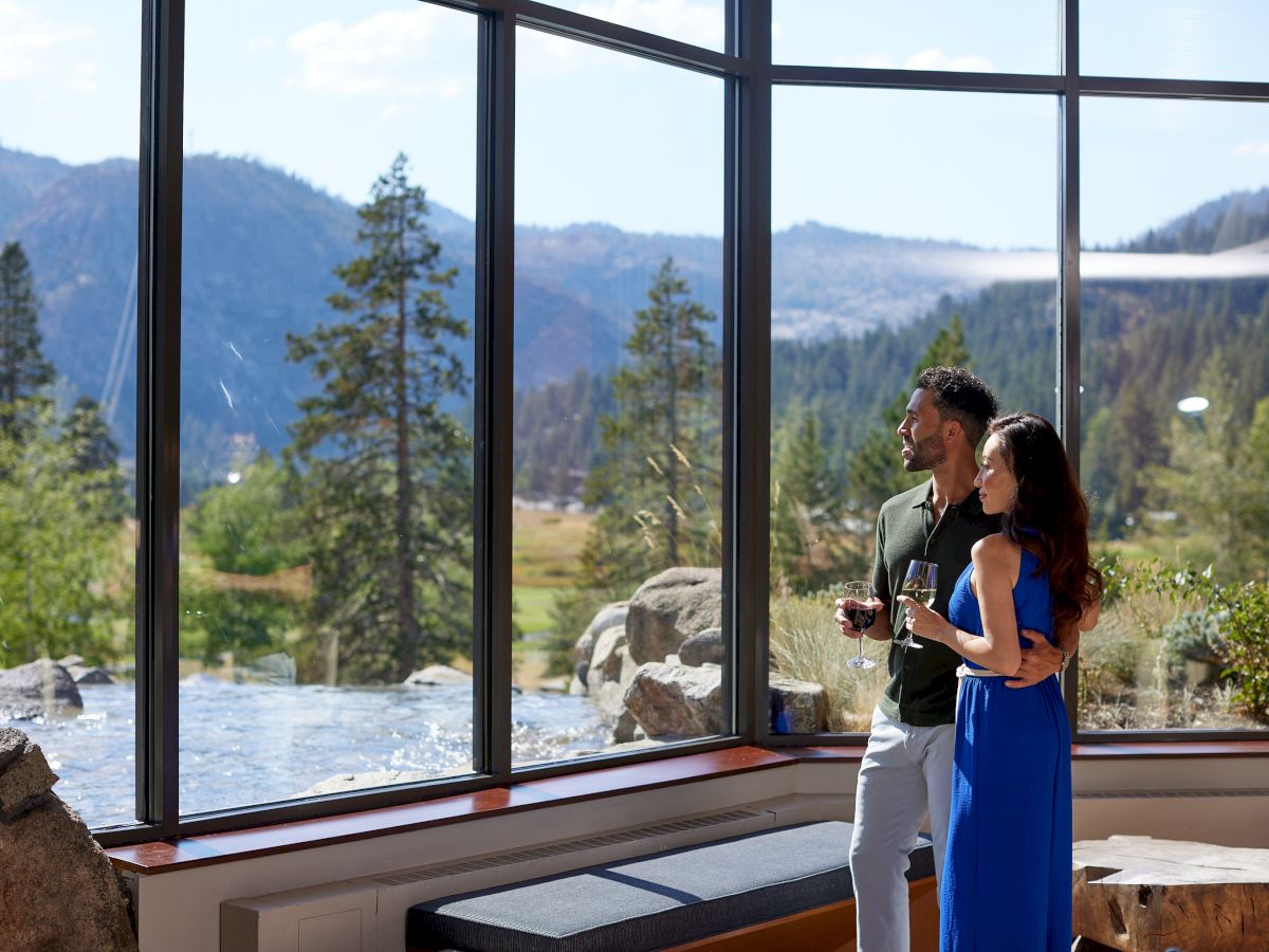 A couple stands by large windows, enjoying wine and a scenic mountain view with trees and clear skies outside.