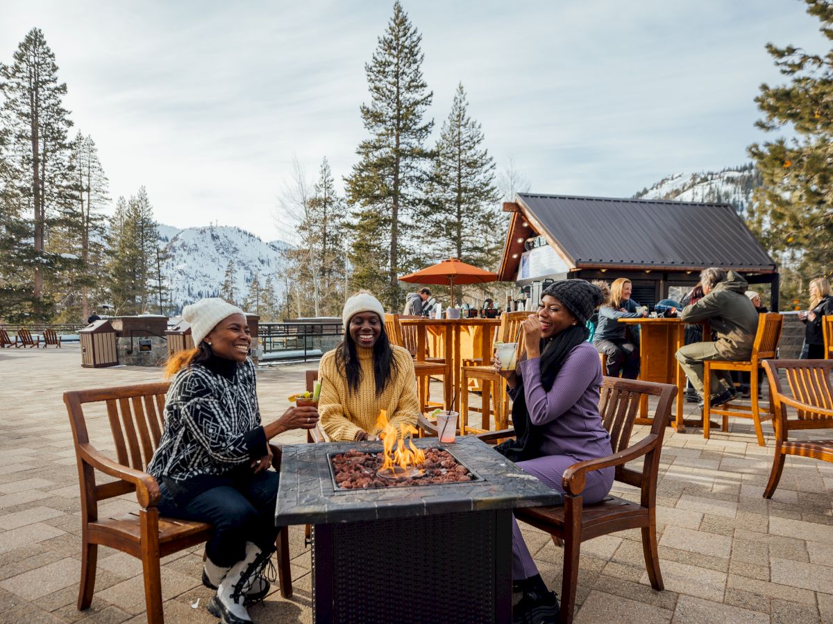 Three people sitting around a fire pit outdoors, wearing winter clothes. Snowy mountains and trees in the background, with a bar nearby.