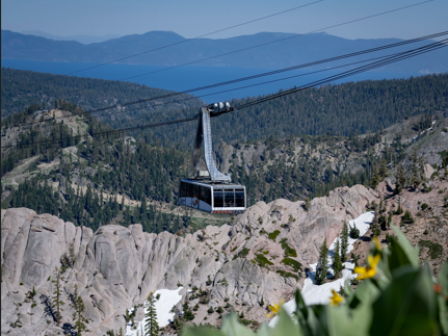 A cable car travels over a mountainous landscape with rocky terrain, trees, and distant mountains under a clear blue sky.