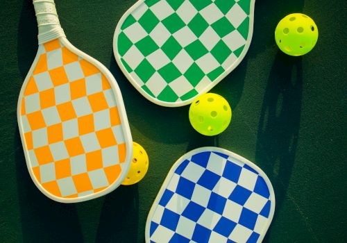The image shows three checkered paddles in orange, green, and blue, alongside three yellow perforated balls on a dark surface.