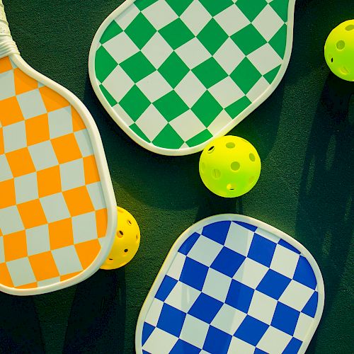 The image shows three checkered paddles in orange, green, and blue, alongside three yellow perforated balls on a dark surface.