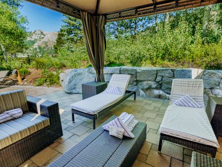 A lounge area with cushioned chairs and chaise lounges on a patio, surrounded by greenery and rocks, under a canopy.