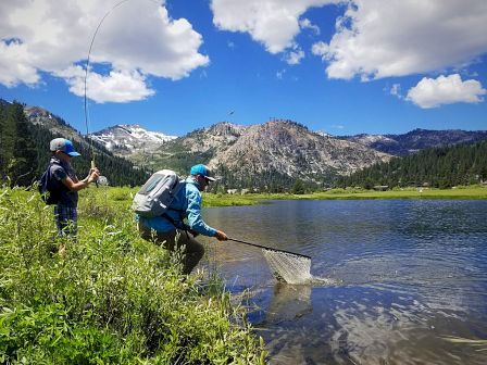Two people are fishing by a scenic lake with mountains in the background, one casting a line, the other with a net.