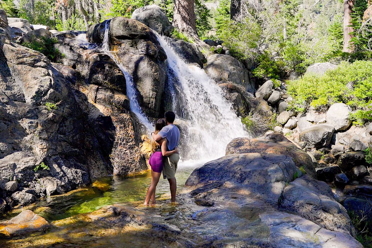 A couple is embracing near a waterfall surrounded by rocks and lush greenery in a forest setting.