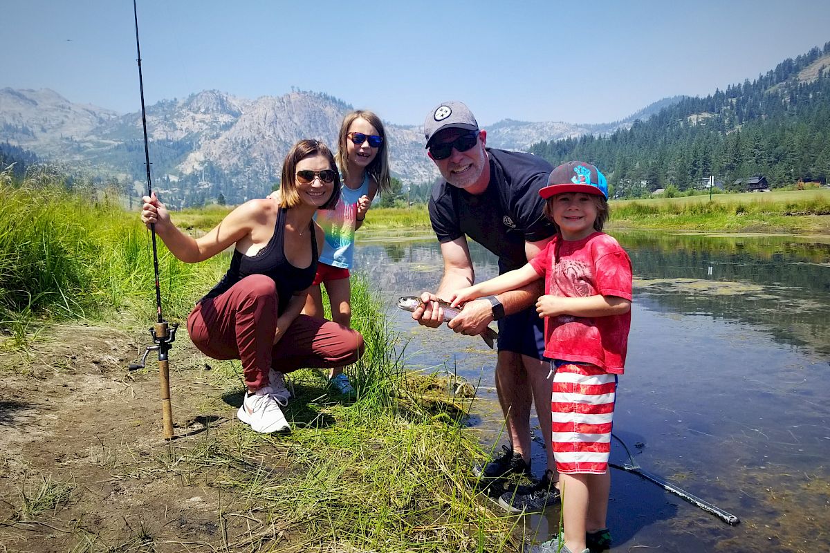 A group of people are posed by the water with fishing gear, surrounded by mountainous scenery and greenery, while smiling at the camera.