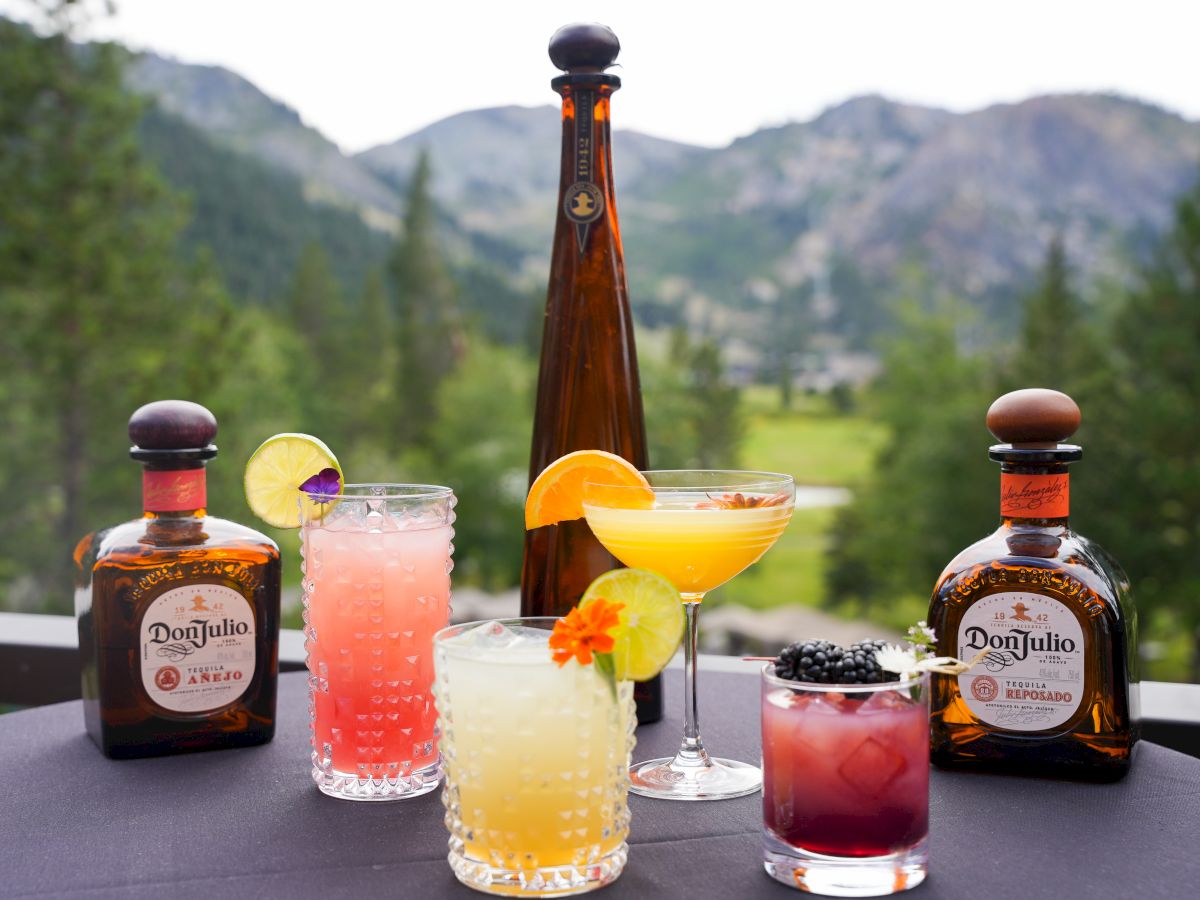 A selection of colorful cocktails and tequila bottles are displayed on a table with a scenic mountain view in the background.
