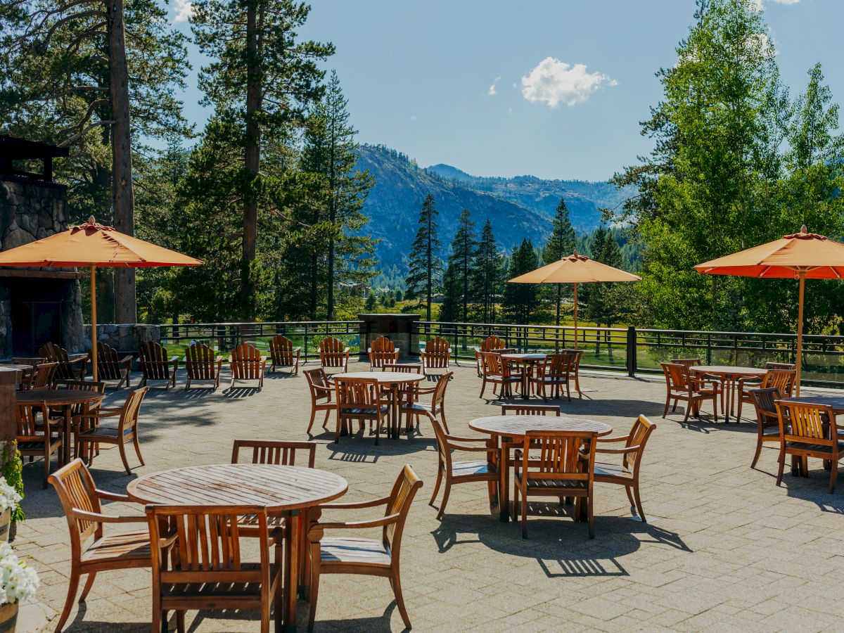 Outdoor patio with wooden tables and chairs, shaded by orange umbrellas, against a backdrop of trees and mountains under a blue sky.