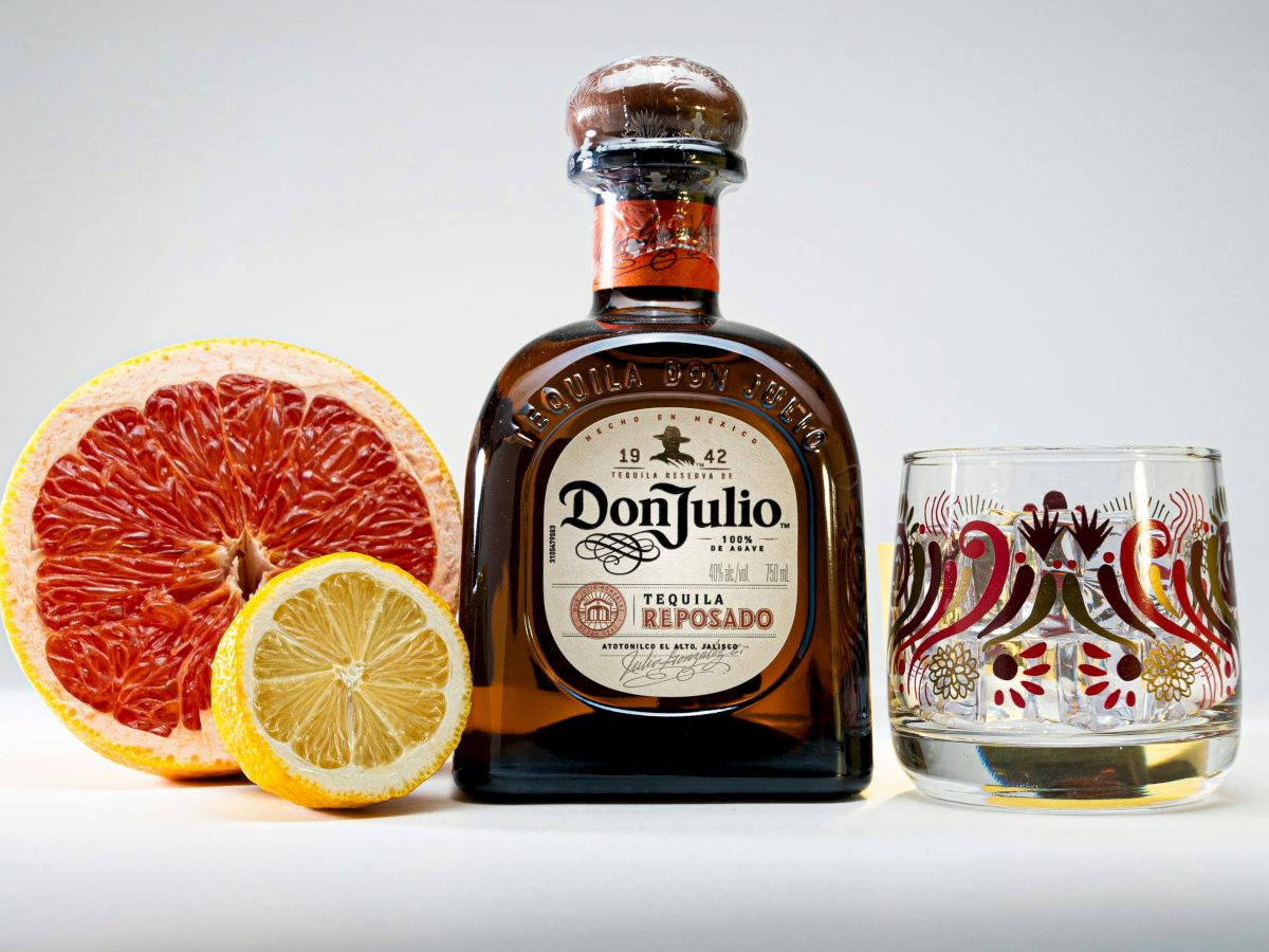 A bottle of Don Julio Reposado tequila is next to half a grapefruit, half a lemon, and an ornate glass on a plain background.