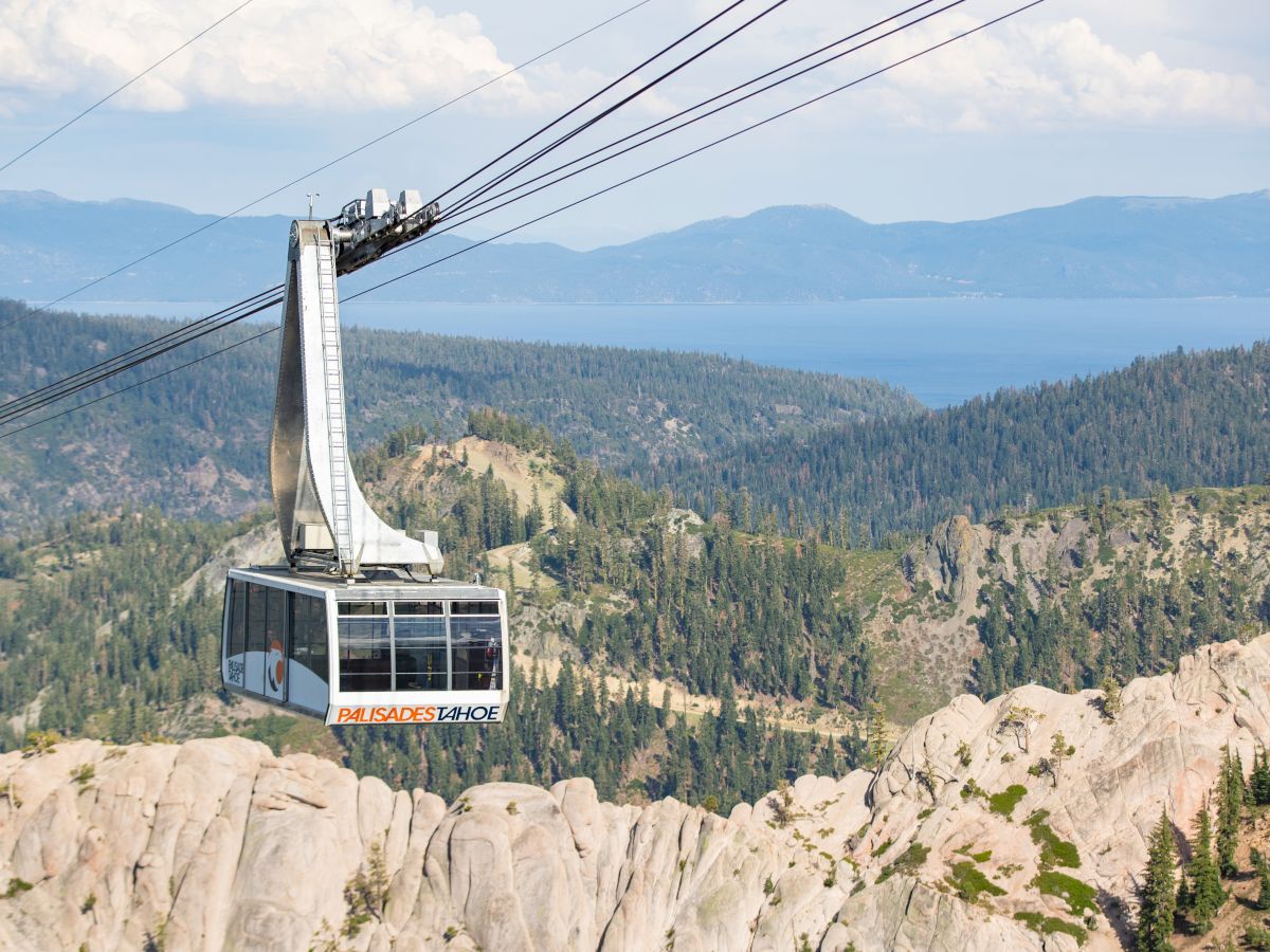A cable car ascends over a mountainous landscape with dense forests and a large lake in the background, under a partly cloudy sky.