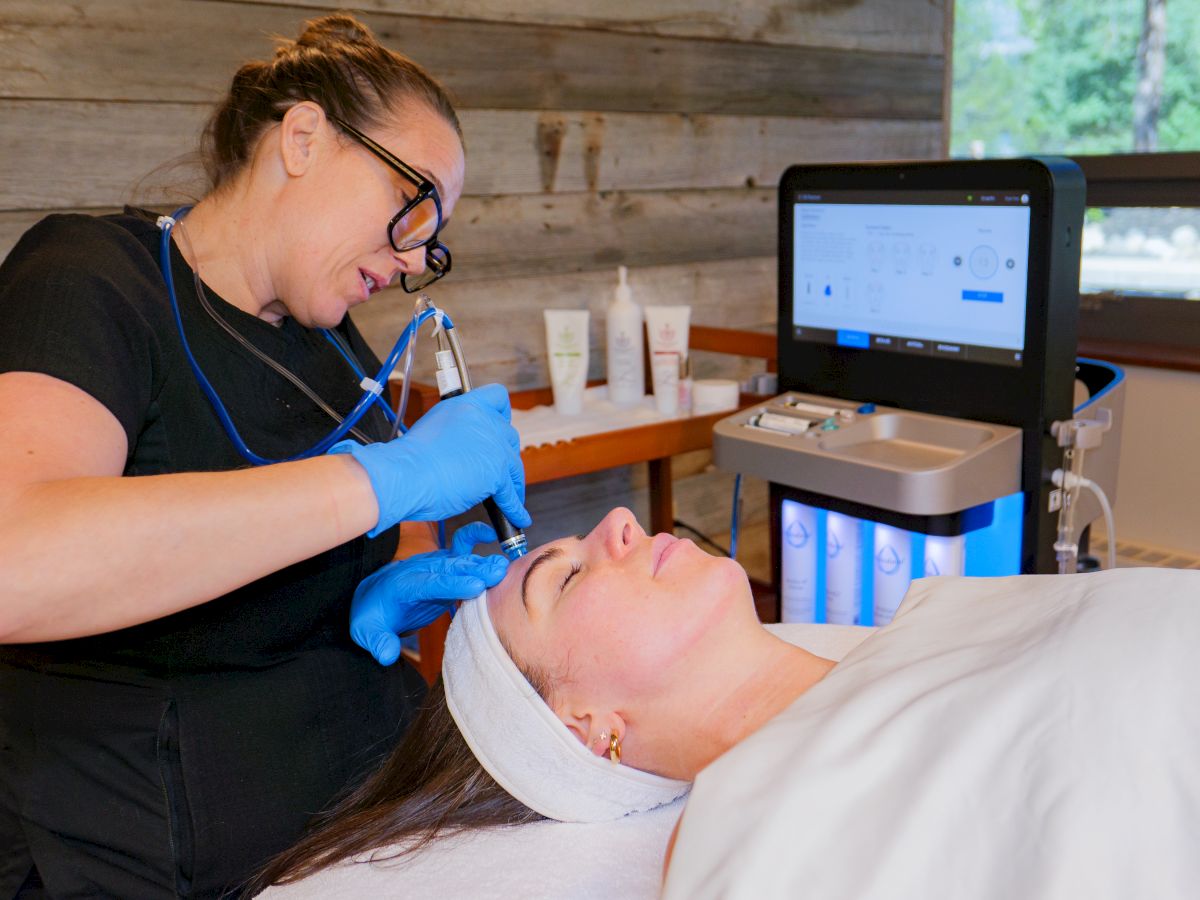 A clinician in blue gloves treats a client&rsquo;s face with a handheld tool while a monitor displays on-screen settings.