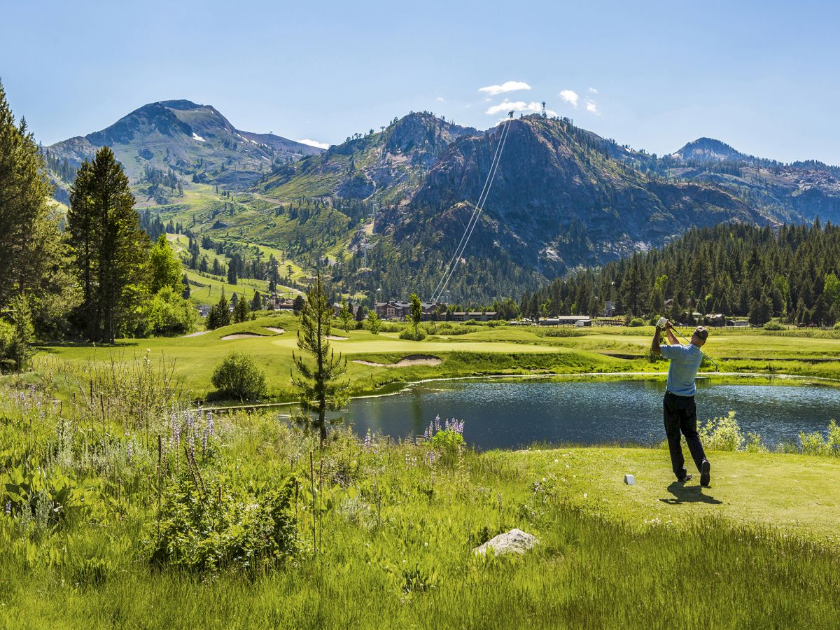 A golfer swings on a lush green course, with mountains and a clear blue sky in the background, near a small pond reflecting the scenery.