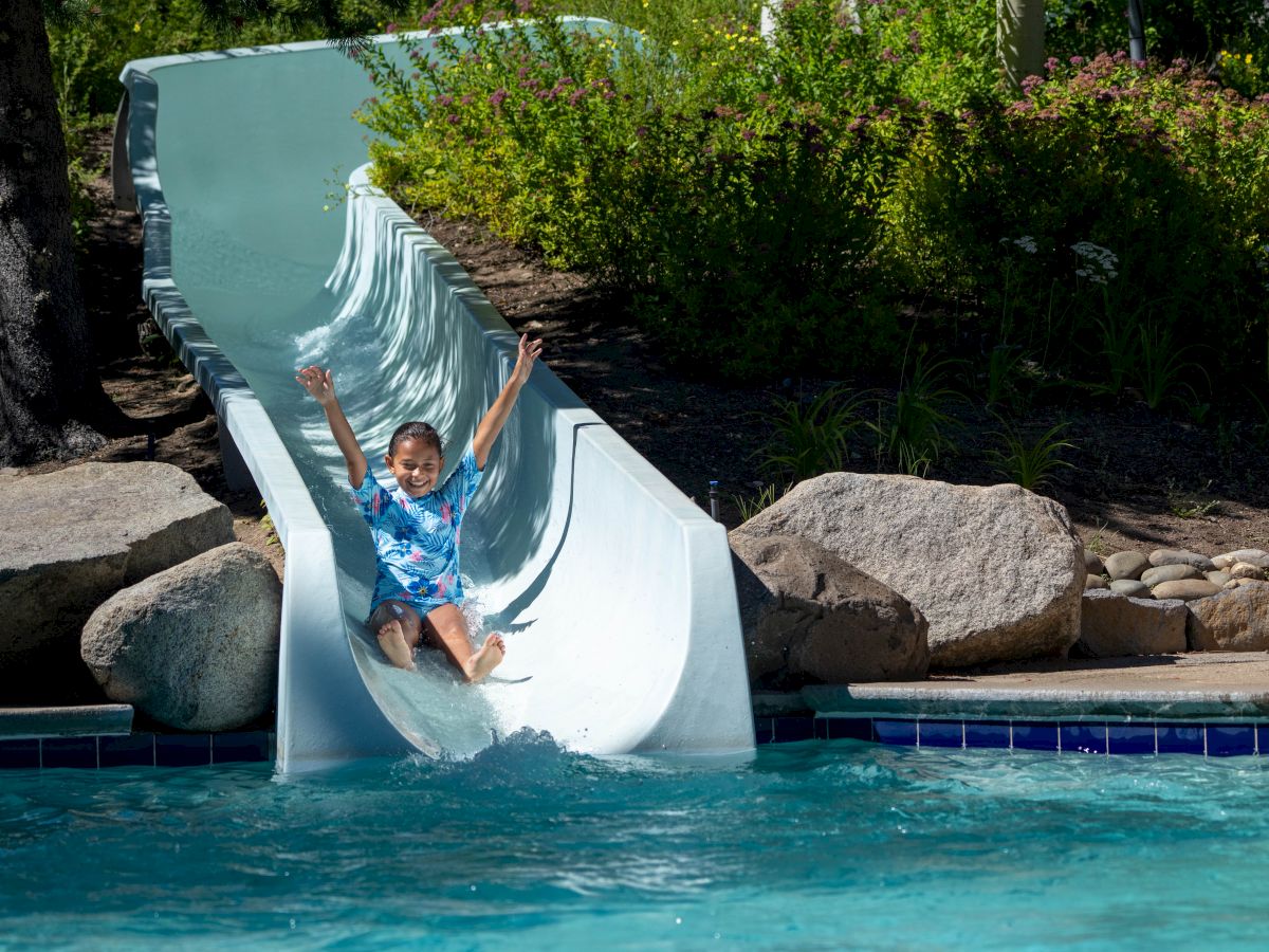 A child in a blue shirt slides down a water slide, arms up, into a pool, surrounded by rocks and greenery.