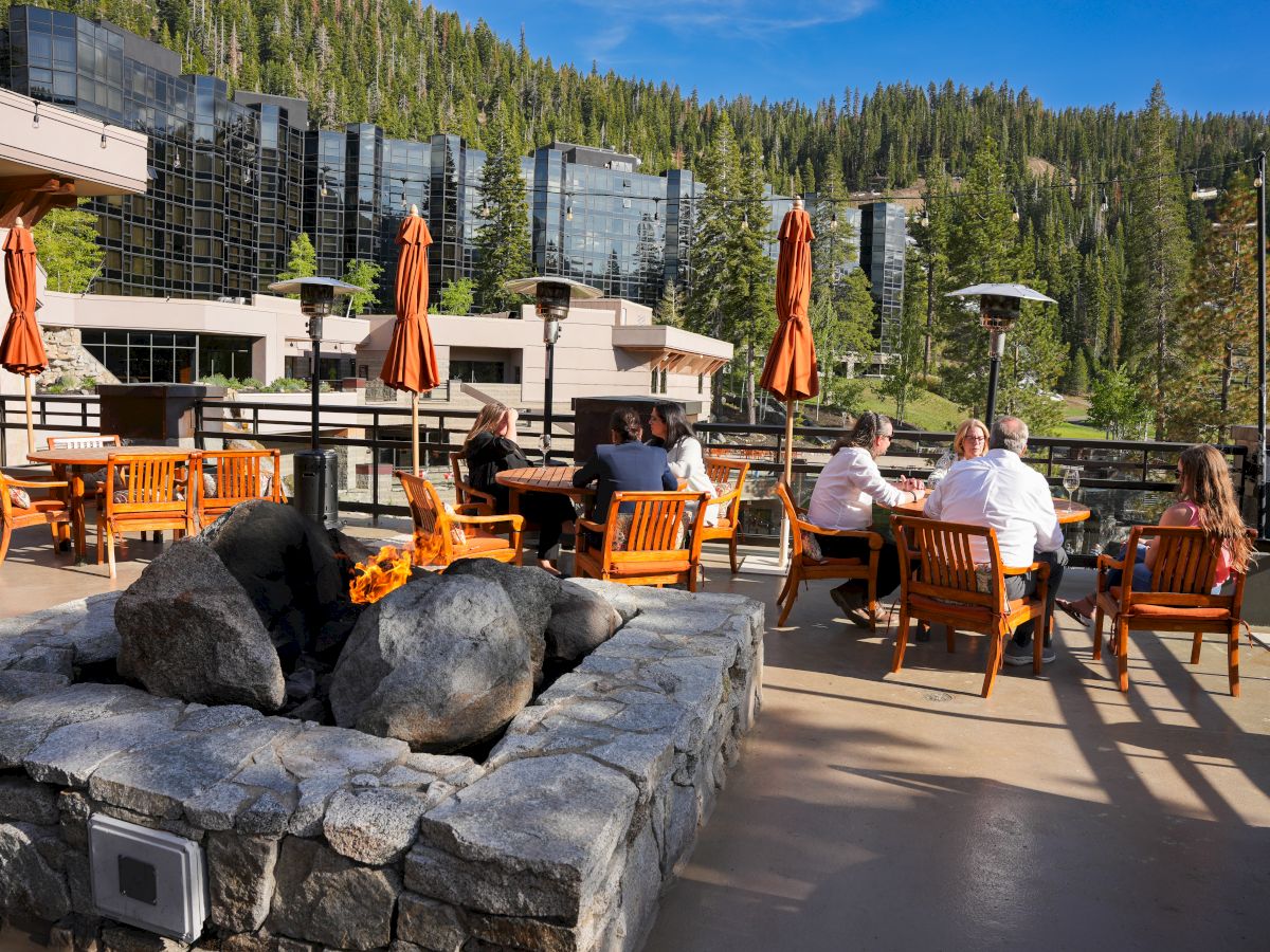 People sitting on a terrace with wooden furniture, surrounded by trees and modern buildings, under a clear blue sky.
