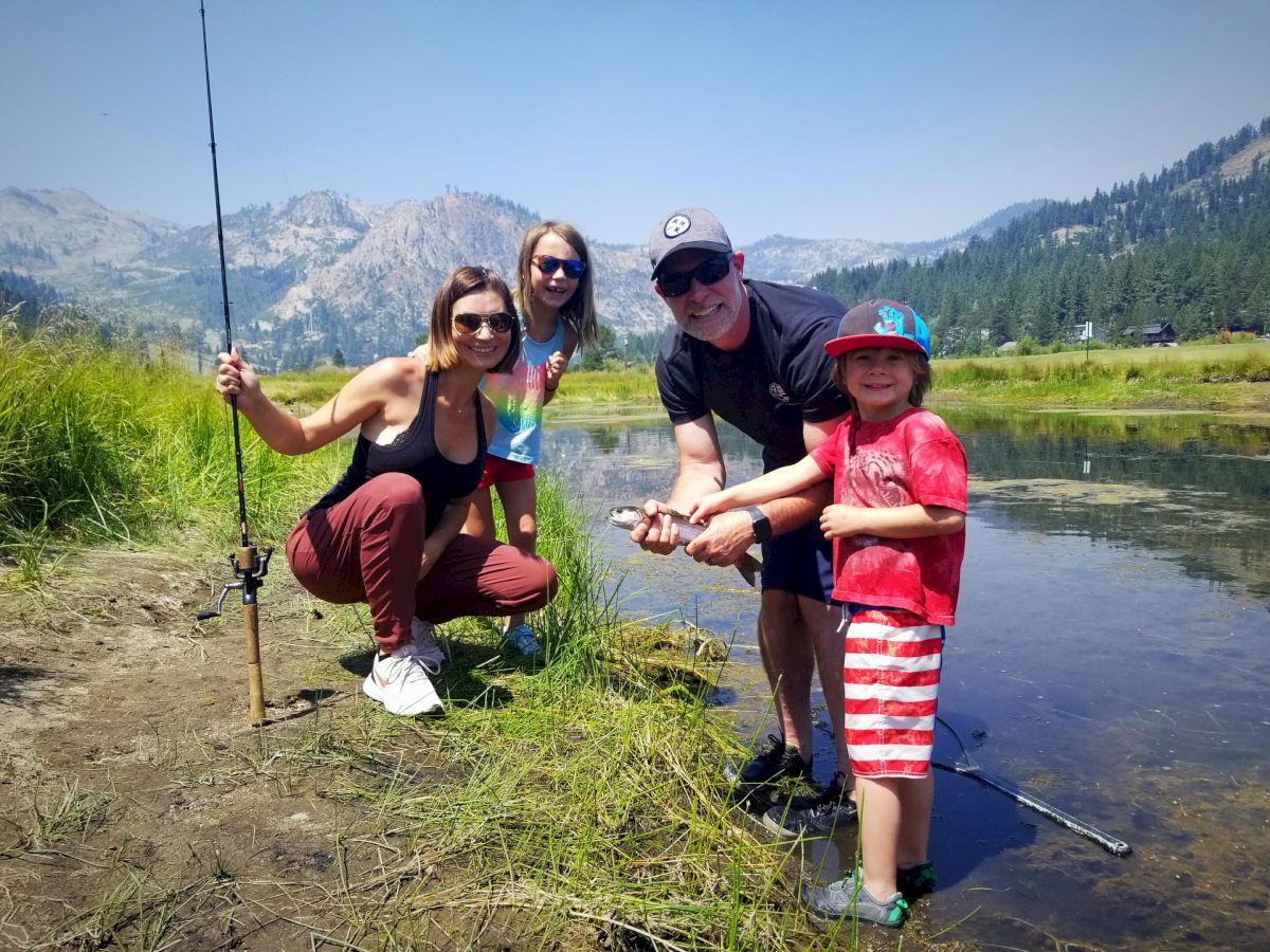 A family of four is fishing by a lake with mountains in the background. They're smiling and holding a fish, enjoying a sunny day together.