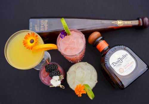 A variety of cocktails garnished with flowers and fruit slices, alongside two bottles of tequila, displayed on a black surface.