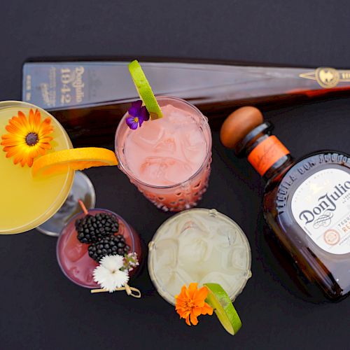 A variety of cocktails garnished with flowers and fruit slices, alongside two bottles of tequila, displayed on a black surface.