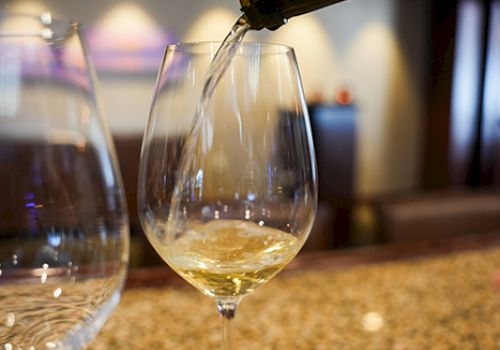 A glass of white wine is being poured from a bottle, with a blurred background and another glass nearby on a countertop.