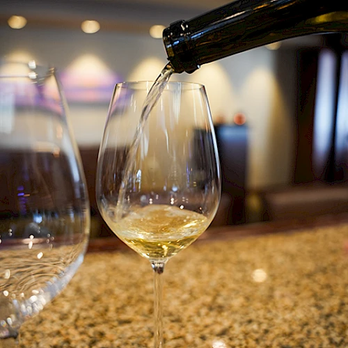 A glass of white wine is being poured from a bottle, with a blurred background and another glass nearby on a countertop.