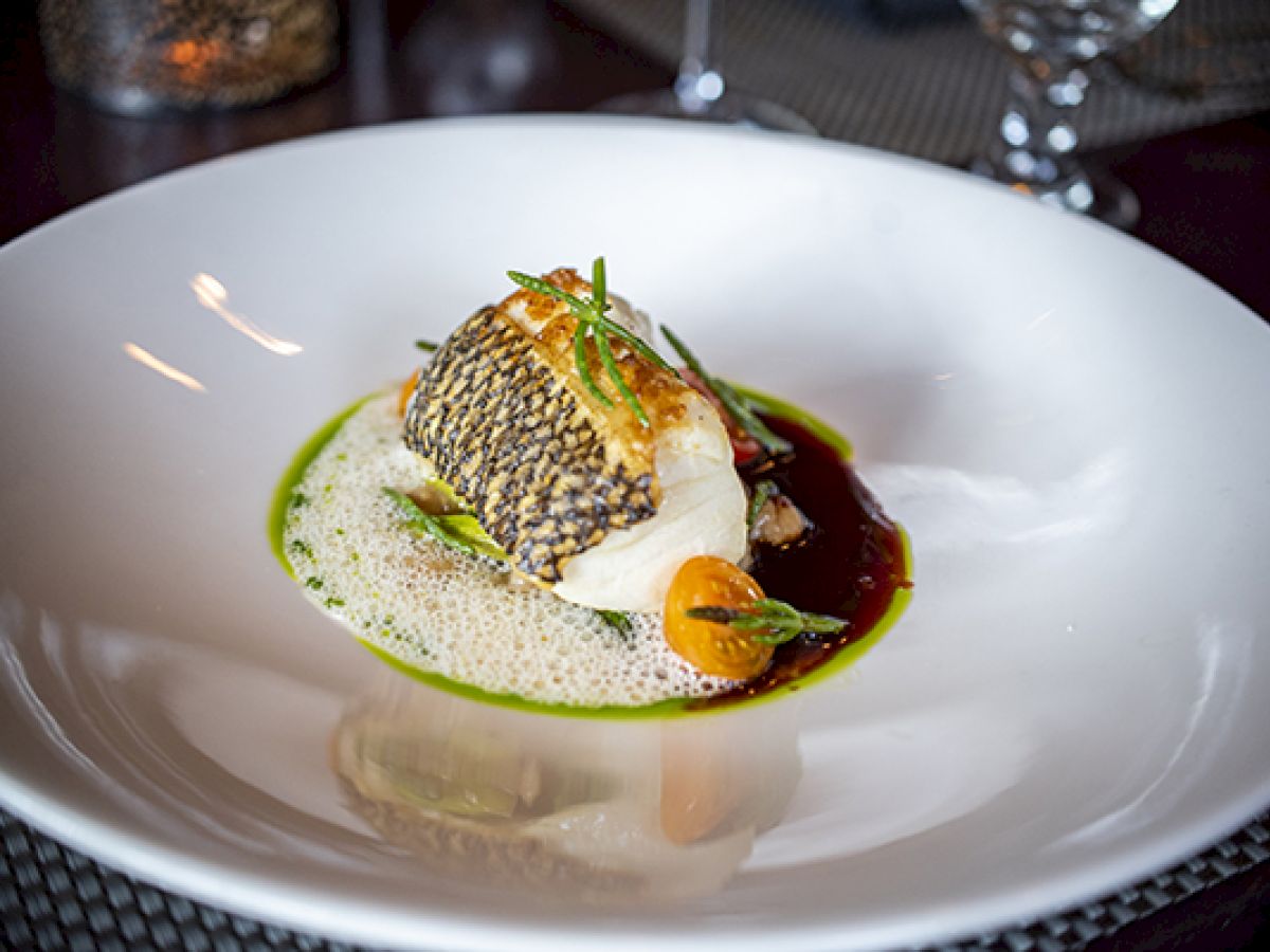 The image shows a plated gourmet dish with seared fish, sauce, foam, and garnishes in a white bowl.