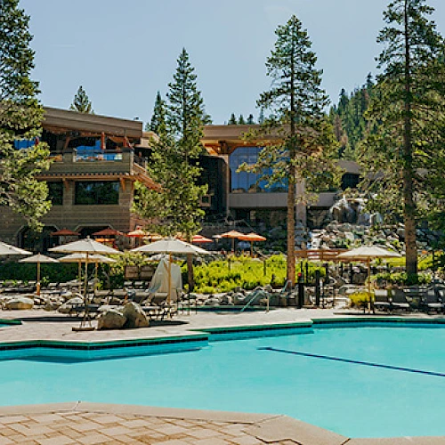 A resort-style pool area with lounge chairs, umbrellas, and a building in the background, surrounded by trees and sunny weather.