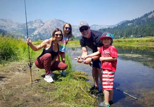 A group of four people by a mountain lake, two hold fishing gear, and one child appears to have caught a fish.