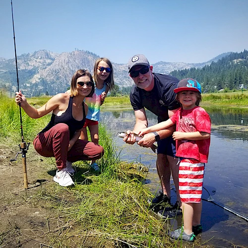 A group of four people by a mountain lake, two hold fishing gear, and one child appears to have caught a fish.