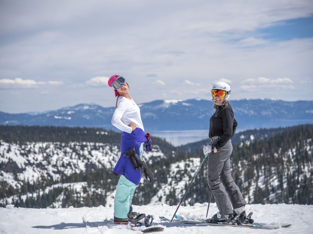 Two people are posing with skiing gear on a snowy mountain, with a scenic view of trees and distant mountains under a blue sky.