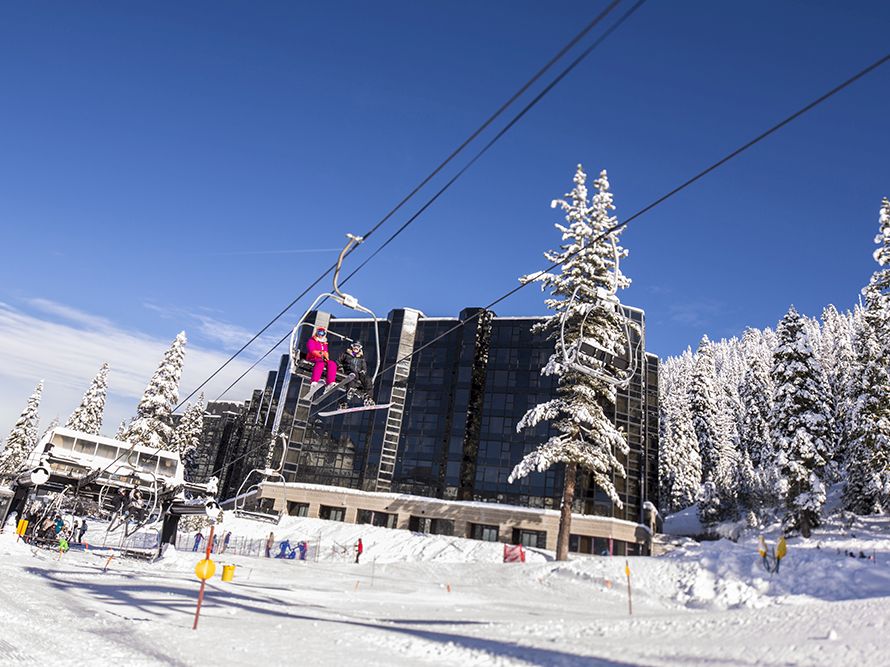 A snowy ski resort scene shows a chairlift with riders passing near a large building and snow-covered pine trees under a blue sky.
