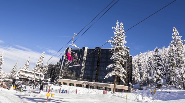 A snowy ski resort scene shows a chairlift with riders passing near a large building and snow-covered pine trees under a blue sky.