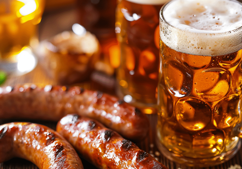 Grilled sausages and frothy beer mugs on a wooden table, creating a cozy, hearty meal scene.