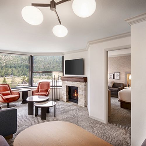 A cozy living room with modern furniture, a fireplace, and large windows showing a green landscape; an adjoining bedroom is visible.