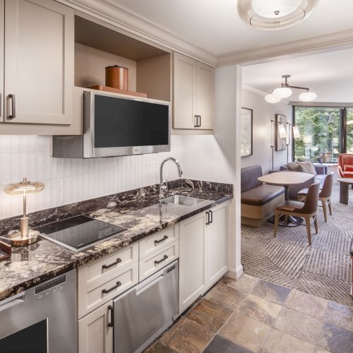 The image shows a modern kitchen and dining area with a marble countertop, built-in appliances, and view into a living area with a large window.