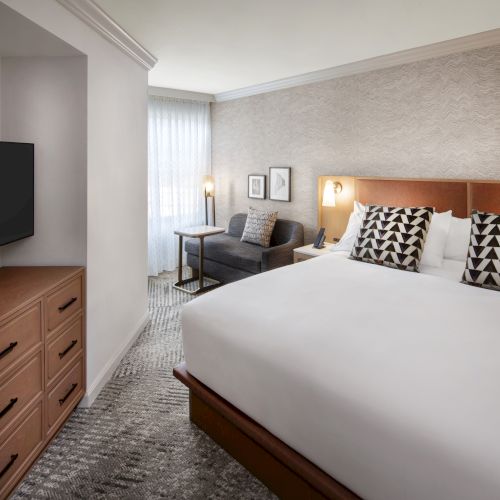 A hotel room with a large bed, geometric patterned pillows, dresser, TV, and a small seating area by the window with natural light.