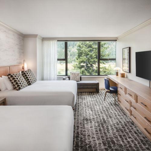 The image shows a hotel room with two beds, a TV, a desk with a chair, and a window with a view of trees outside.