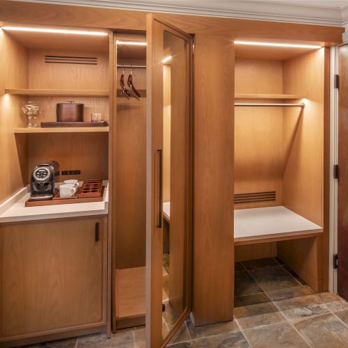 Cozy wooden closet with shelves, a coffee maker, cups, and a hanger bar, next to a closed wooden door in a room.