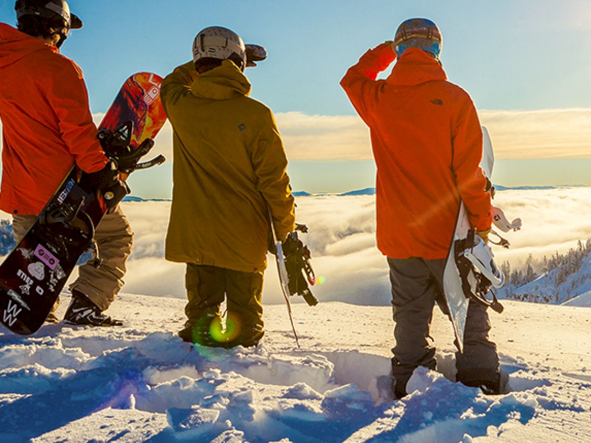Three people in winter gear with snowboards stand on a snowy peak, gazing at the bright, sunny horizon above the clouds.