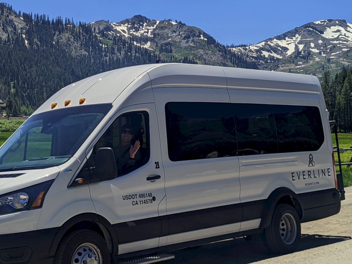 The image shows a white passenger van parked on a road with mountainous scenery and snow-capped peaks in the background, under a clear blue sky.