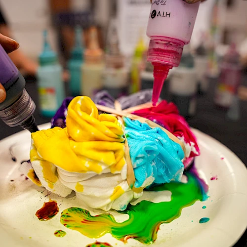 A colorful pile of frosting blobs in yellow, blue, green, and pink being squeezed onto a white plate, creating a messy, rainbow paint-like dessert.