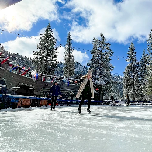 Two people skate on a sunny outdoor ice rink tucked among snow-covered trees, with colorful seats and flags along the side.