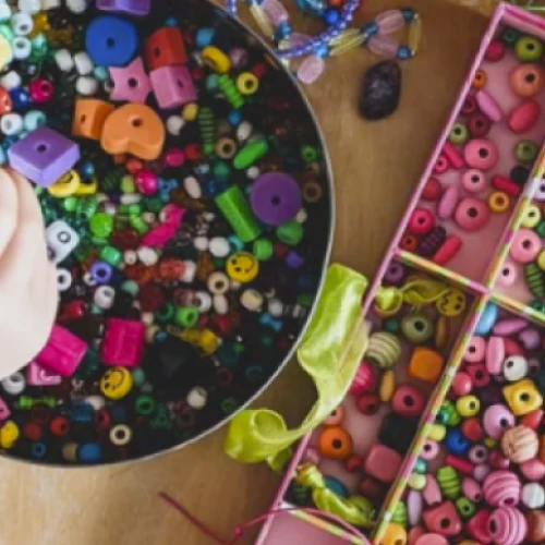 A colorful craft scene with a hand sorting plastic beads and alphabet letters over a bowl of mixed beads and separate bead organizers on a table.