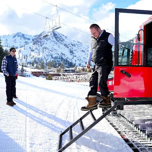 Two people loading a snowboarder onto a red snowcat for a ride in a snowy mountain setting.
