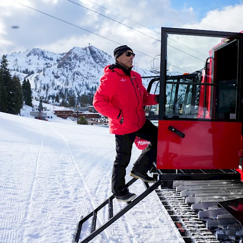 A man in a red jacket and black pants steps off a red snowcat onto a snowy slope with mountains in the background, preparing for a ride.