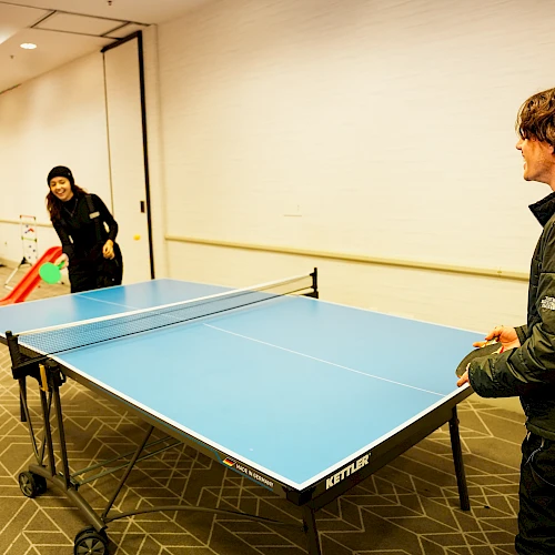 Two people play table tennis in a room; one prepares a serve while the other watches near the net.